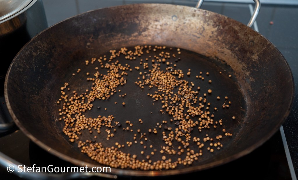 A cast iron pan with toasted coriander seeds scattered across the bottom, showcasing the spices used in cooking.