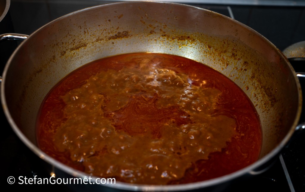A close-up of a pot filled with a bubbling, rich curry broth, showcasing a deep reddish-brown color and a textured surface.
