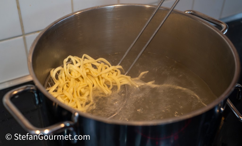 Fresh egg noodles being cooked in boiling water, with a pair of tongs holding some of the noodles.
