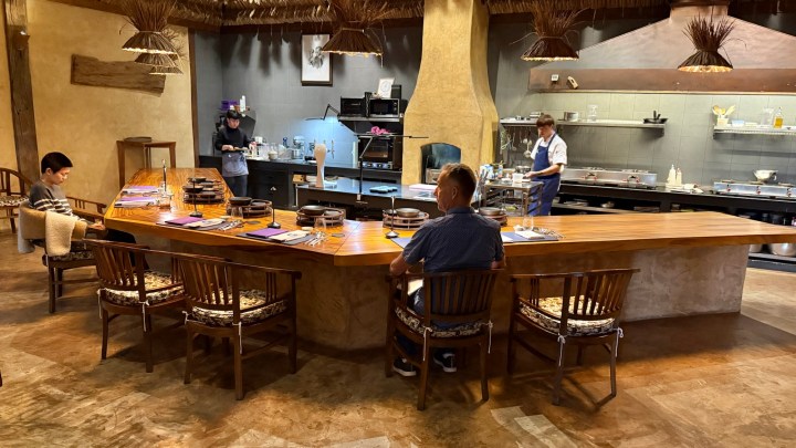 Interior view of a chef's table restaurant featuring a wooden counter with guests seated, an open kitchen area with chefs preparing food in the background.
