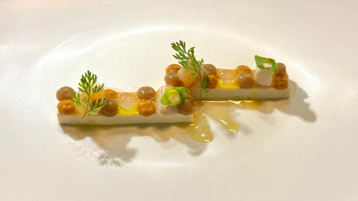 An elegant dish featuring rectangular pieces of soft white tofu or a similar ingredient, topped with small, round golden-brown elements, a drizzle of sauce, and garnished with green herbs and scallions.