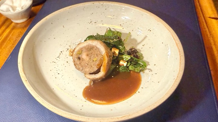 A plated dish featuring a sausage roll made of pork belly filled with Lanna Larb, garnished with greens and accompanied by a savory sauce.