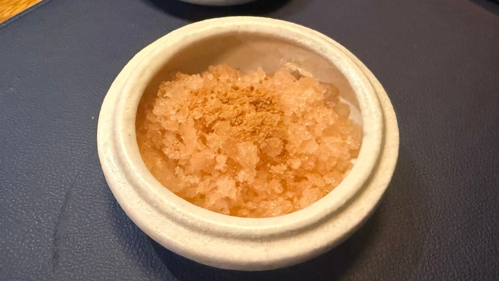 A bowl containing a pink granita with a sprinkle of brown powder on top, placed on a dark blue surface.