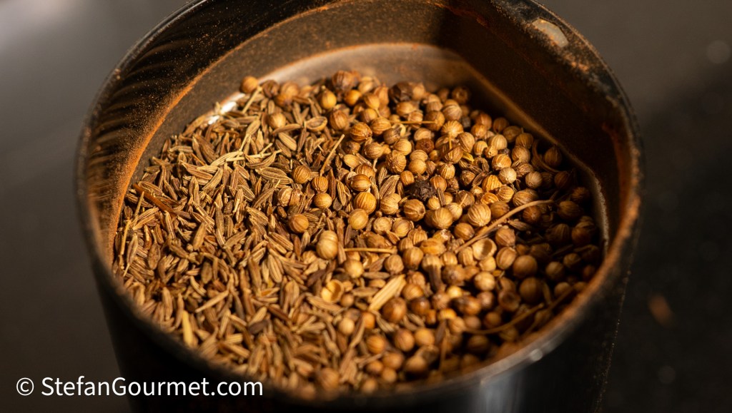 A close-up of a mix of toasted spices, including coriander seeds and cumin, in a black container.