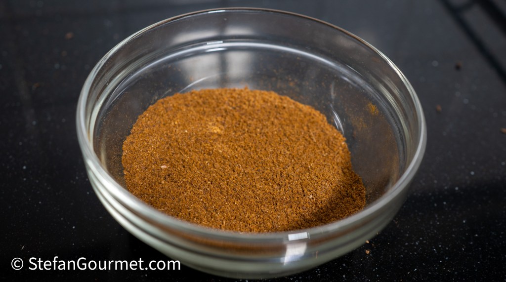 A small glass bowl filled with a fine brown spice blend for Gaeng Hung Lay curry, set on a dark surface.