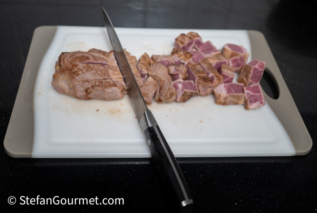 Varkensbuik wordt met een mes in plakjes gesneden op een snijplank, waarbij zowel de hele als de in blokjes gesneden stukken zichtbaar zijn.