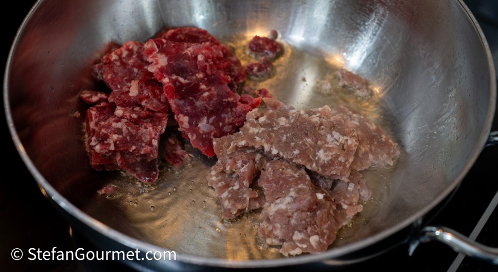 A pan with a mixture of coarsely ground beef and sweet Italian sausage cooking over heat, showing the meat sizzling as it browns.
