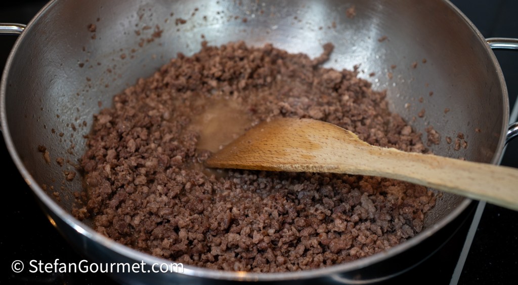 A pot with coarsely ground beef cooking, stirred with a wooden spoon, releasing juices as it browns.