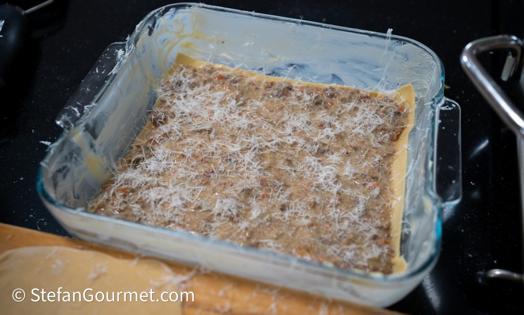 A glass baking dish with layers of fresh pasta and a creamy meat sauce, topped with grated cheese, prepared for baking.