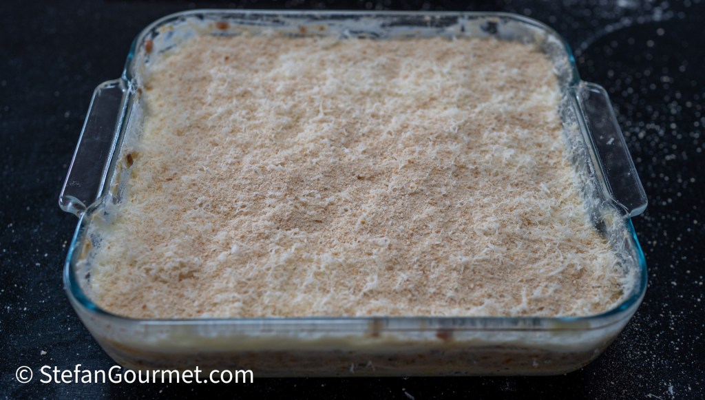 A glass dish containing freshly prepared lasagna topped with a layer of breadcrumbs and Parmesan cheese, ready to be baked.