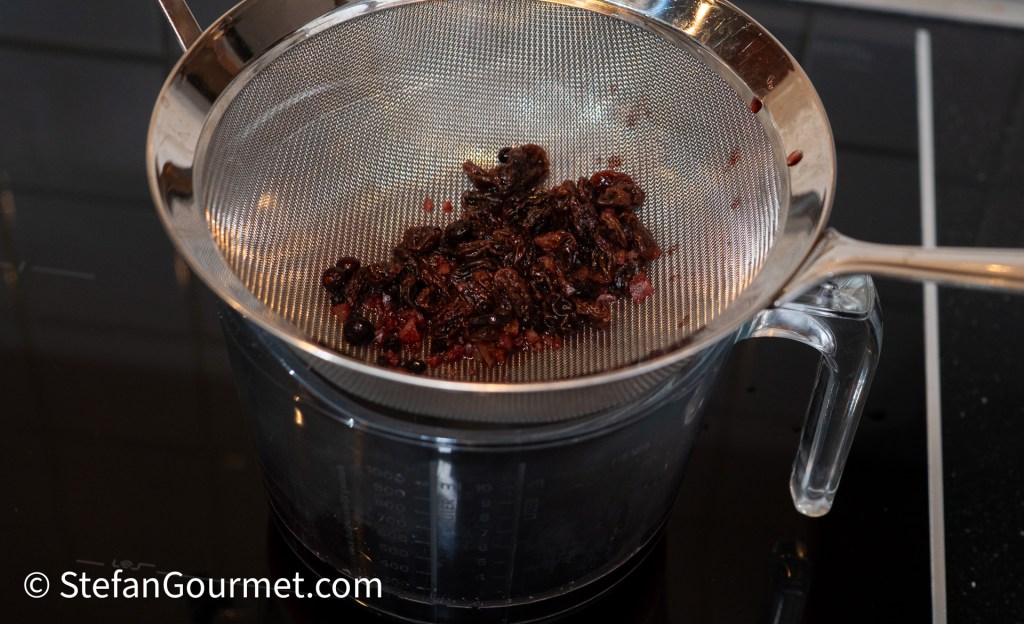 A strainer holding soaked raisins and aromatic ingredients over a measuring jug, showcasing the preparation process for a gourmet sauce.