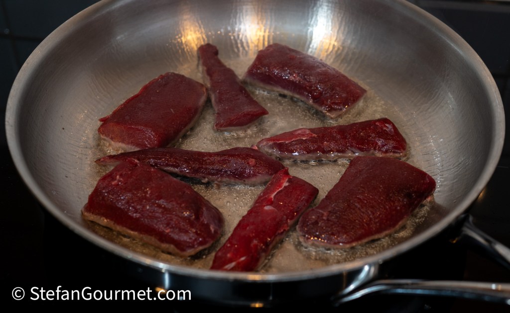 Several pieces of hare fillet searing in a stainless steel pan with a light browning and some fat that has melted.