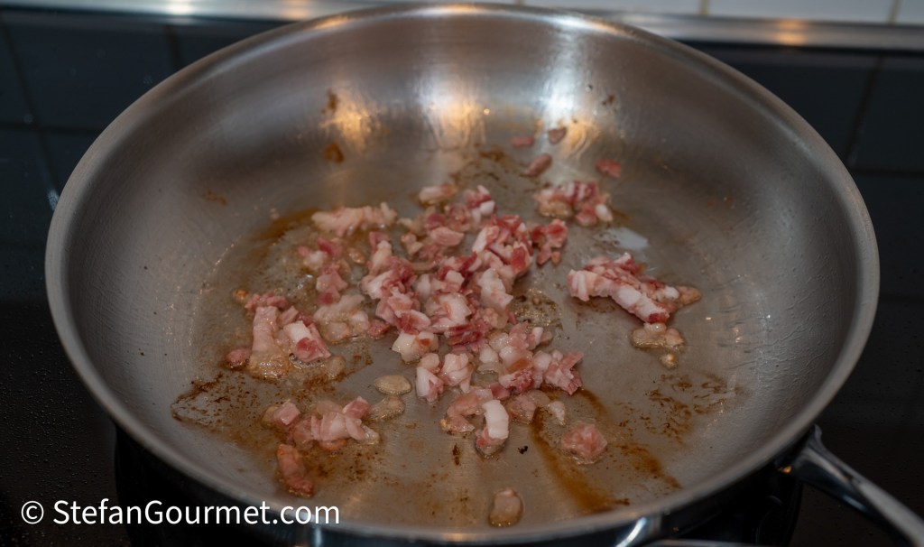 Diced pancetta cooking in a stainless steel frying pan, releasing fat and browning.