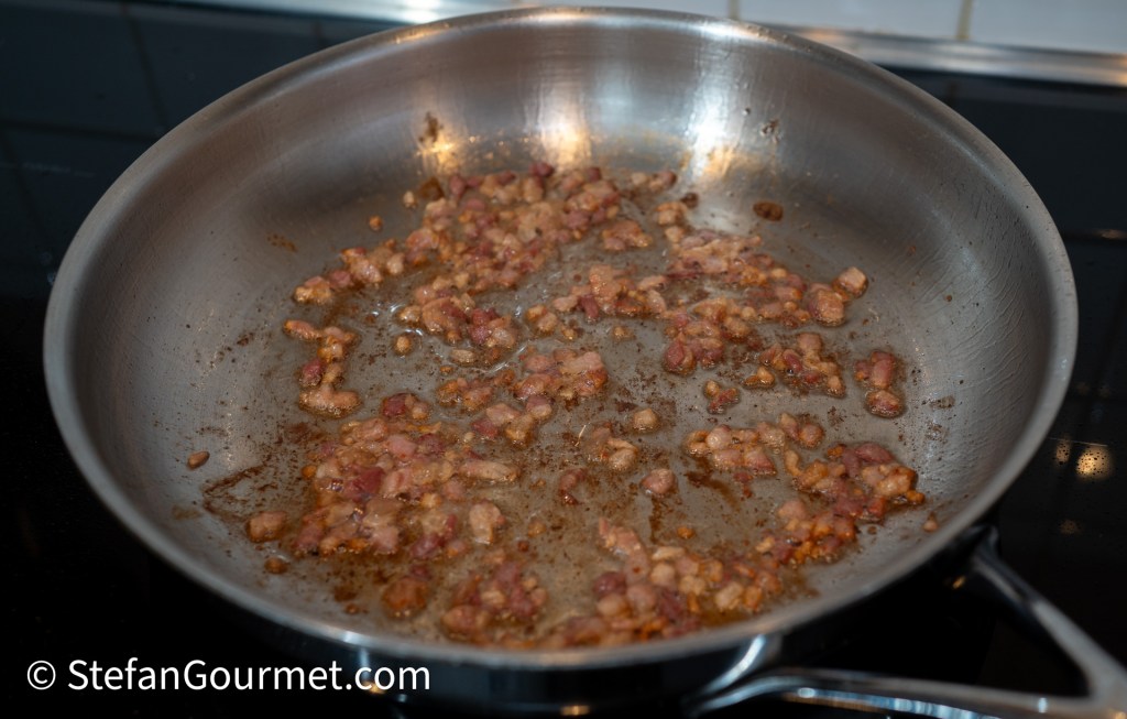 Browning diced pancetta in a stainless steel frying pan on the stove, with some crispy bits scattered on the bottom.