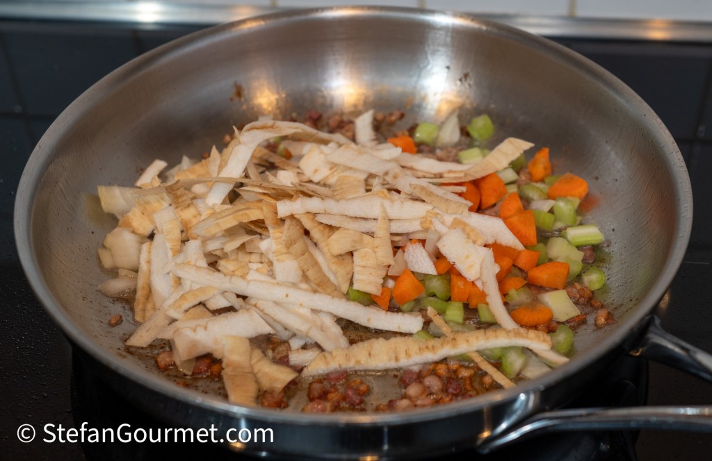 A pan with sautéing vegetables, including strips of parsley root, diced carrots, and celery, on a stovetop.