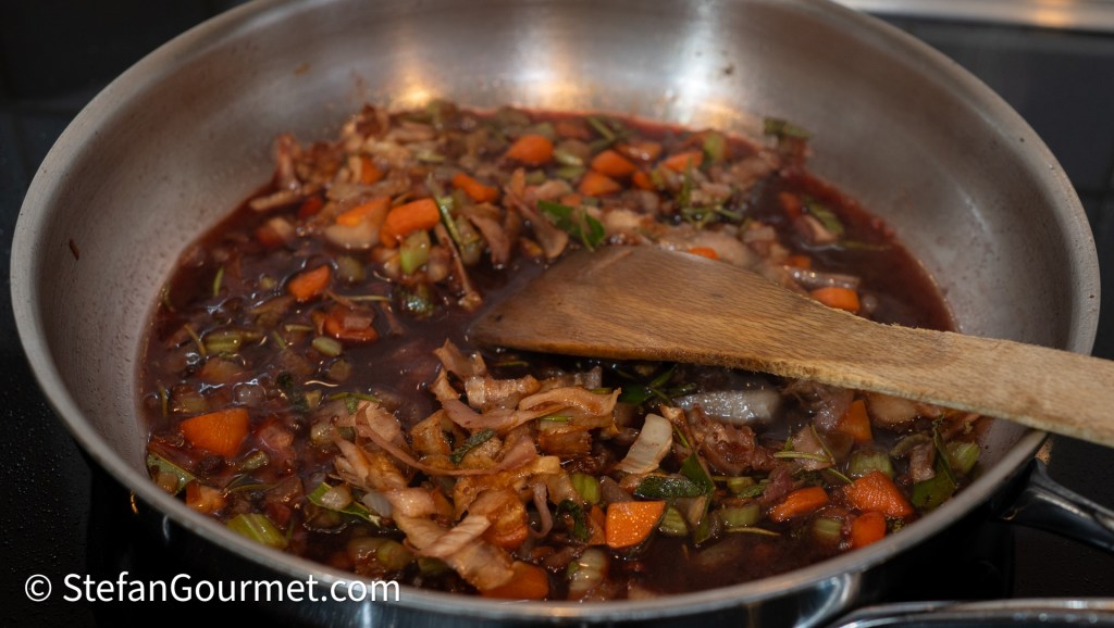 A saucepan containing a dark red sauce with chopped vegetables, including carrots and celery, and herbs being stirred with a wooden spoon.