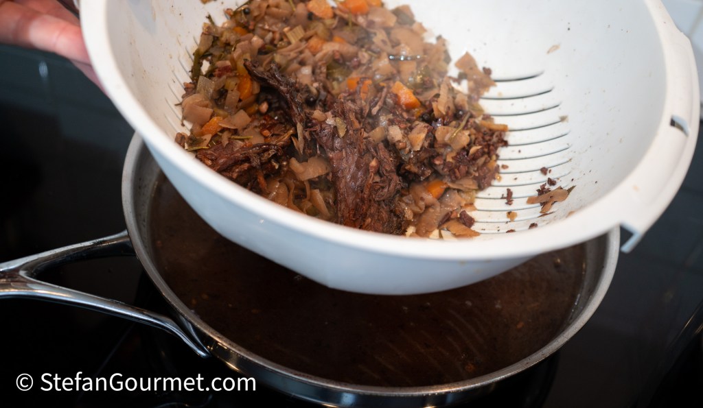Pouring strained vegetables from a colander into a pot containing dark sauce, with steam rising from the mixture.