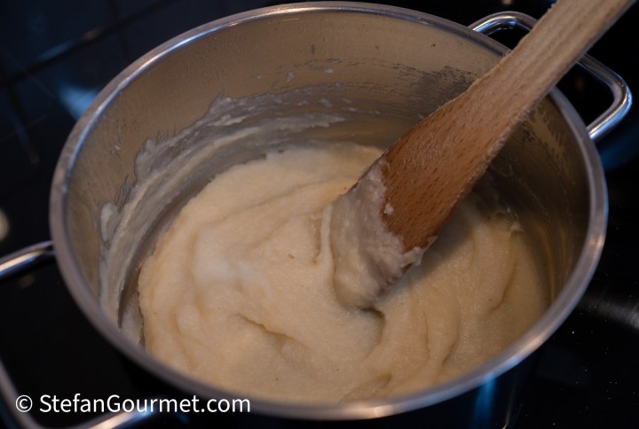 A pot containing a smooth, creamy parsley root puree being stirred with a wooden spoon.