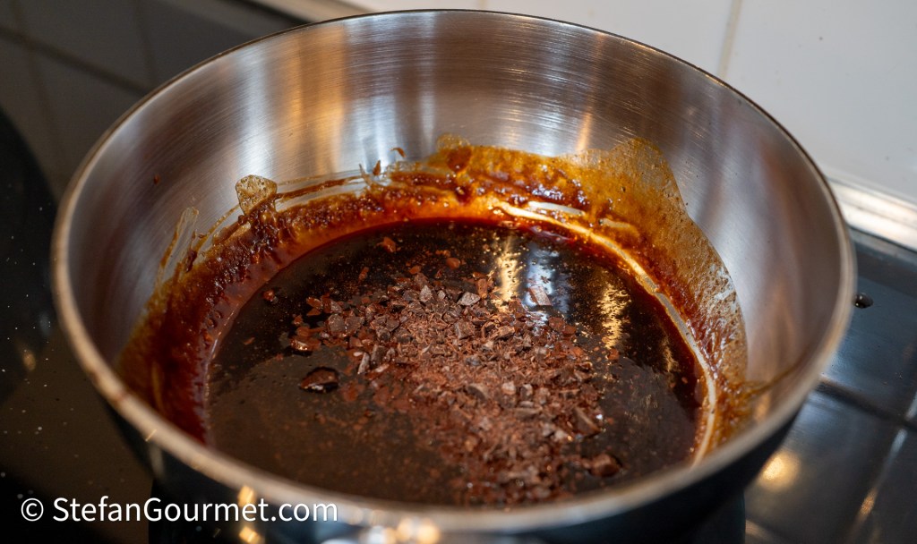 A stainless steel saucepan with a dark sauce and small pieces of minced dark chocolate melting in the bottom.
