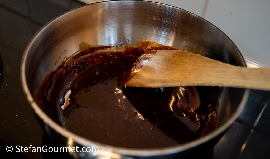 A wooden spoon stirring a thick, dark sauce in a stainless steel saucepan on a stovetop.