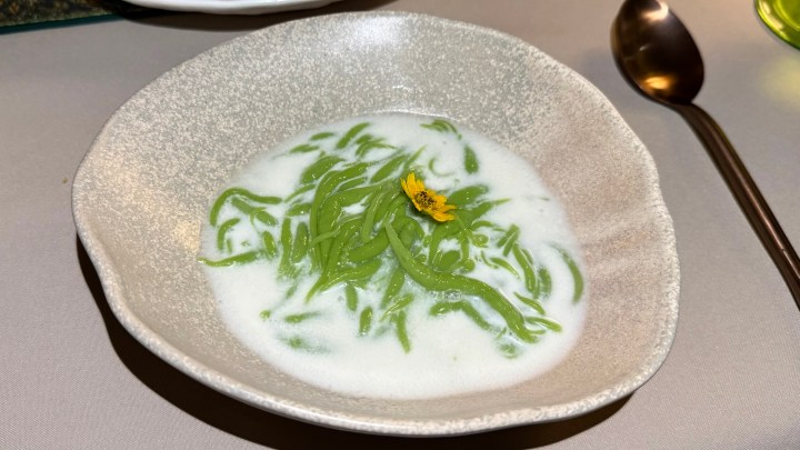 A serving of Lod Chong Nam Kati, featuring pandan jelly noodles in coconut milk, garnished with a yellow flower, presented in a textured bowl.