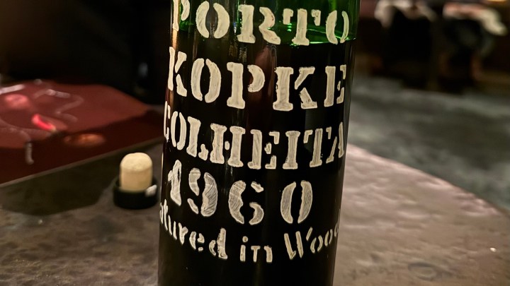 Close-up of a bottle of 1960 Kopke Colheita port wine, featuring white text on a dark background, with a cork and blurred restaurant ambiance in the background.