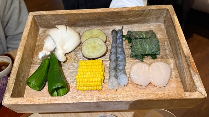 A wooden tray displaying an assortment of fresh ingredients for tempura, including green peppers, corn, sweet potato, shrimp, shiso leaf-wrapped squid, and scallops.