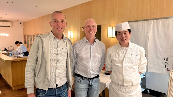 Three men posing together in a modern Japanese restaurant, with a warm wooden interior and soft lighting.