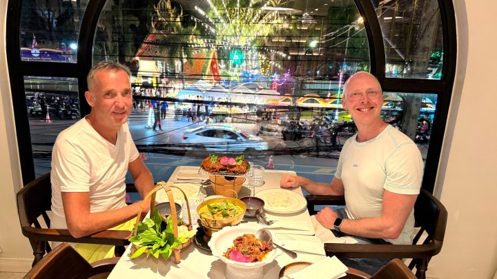 Two men sitting at a restaurant table with a view of a busy street outside, enjoying a meal with various Thai dishes displayed.