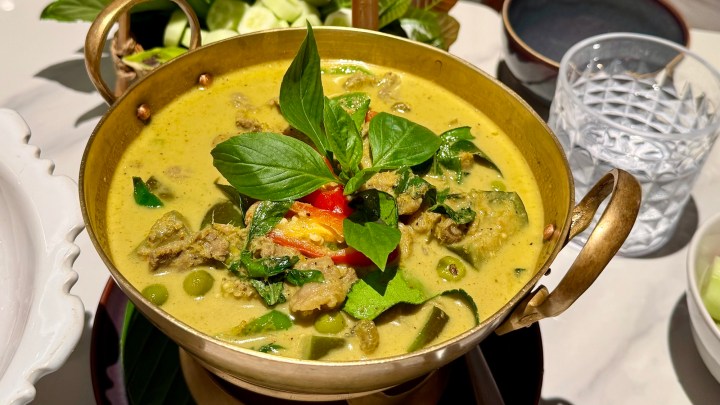 A bowl of green curry with beef, garnished with fresh basil, green peas, and red bell pepper served in a traditional brass pot.