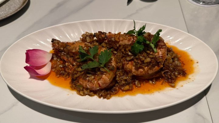 A plate of grilled shrimp served with a flavorful sauce, garnished with herbs and a lotus flower petal.