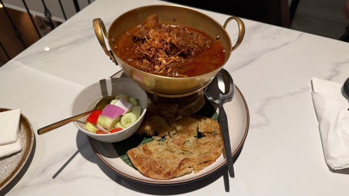 A bowl of green curry with beef topped with crispy onions, served with a side of roti and a small dish of pickled vegetables on a white plate.