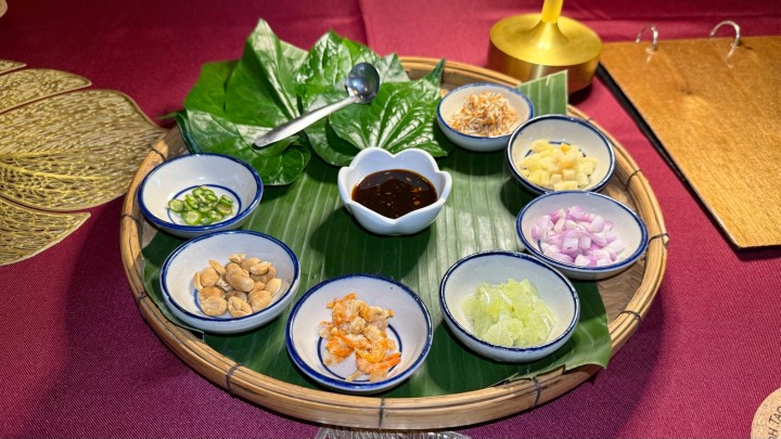 A tray featuring various ingredients for Miang Kham, including betel leaves, a sauce, and assorted toppings like peanuts, dried shrimp, and herbs.