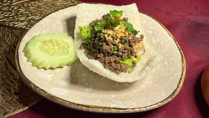 A serving of Laap Kua, a Northern Thai dish featuring a mixture of minced beef, herbs, and spices, presented on a crispy rice cracker, garnished with fresh vegetables and served with a cucumber slice.