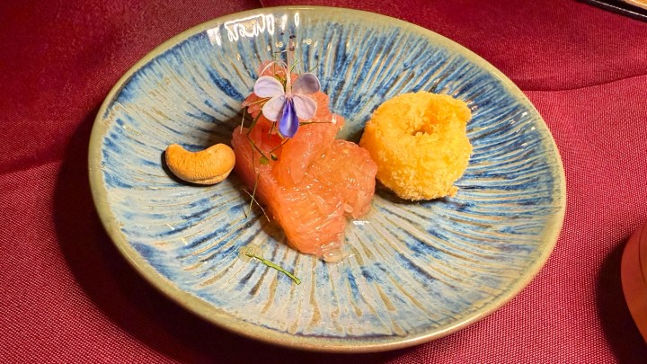 A beautifully presented dish featuring sliced raw fish topped with a delicate flower garnish, served alongside a yellow ball of what appears to be a savory or sweet accompaniment, with a cashew nut on the side, all arranged on a textured blue plate.