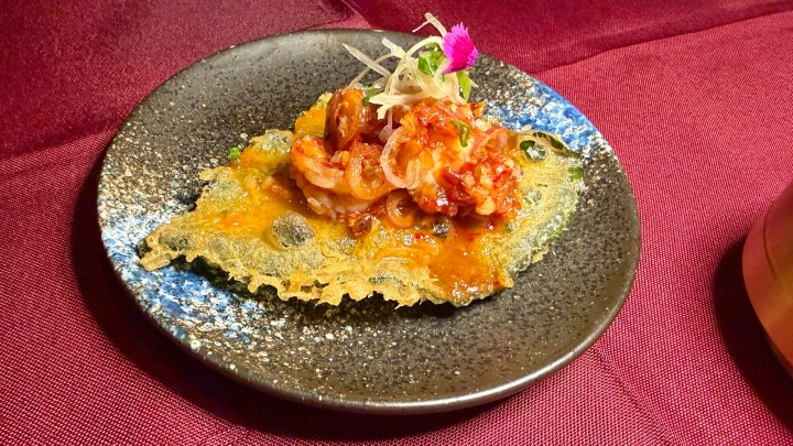 A plate featuring a crispy fried mulberry leaf topped with shrimp salad, garnished with sliced onions and a small flower.