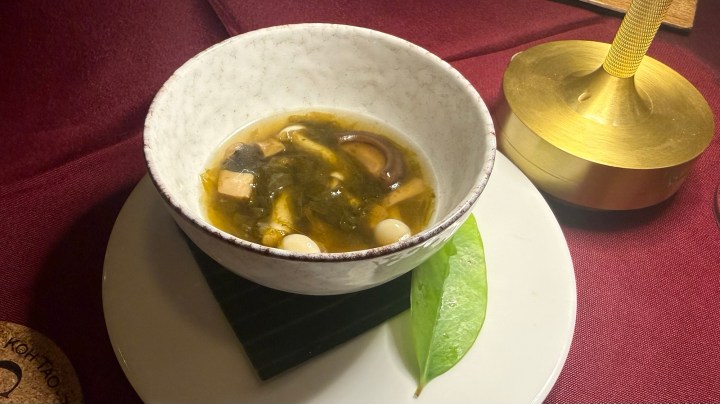 A bowl of traditional Thai soup with assorted mushrooms and herbs, served on a black slate platter, accompanied by a green leaf decoration.