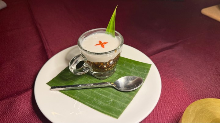 A small glass filled with a layered dessert, topped with coconut cream and a red flower garnish, presented on a banana leaf over a white plate.