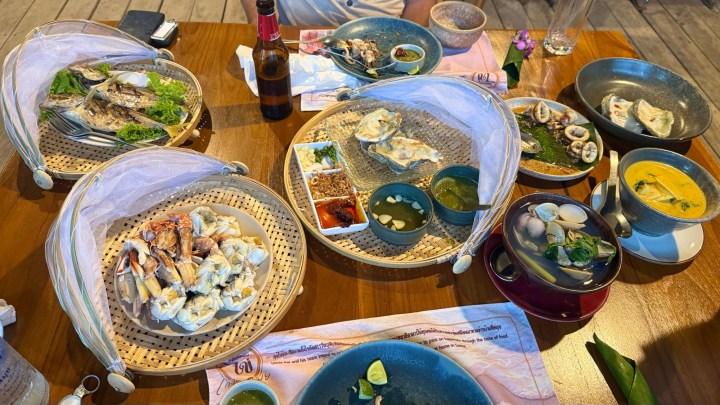A spread of Southern Thai seafood dishes on a wooden table, featuring various ingredients and presentations, with protective mesh covers over some plates.