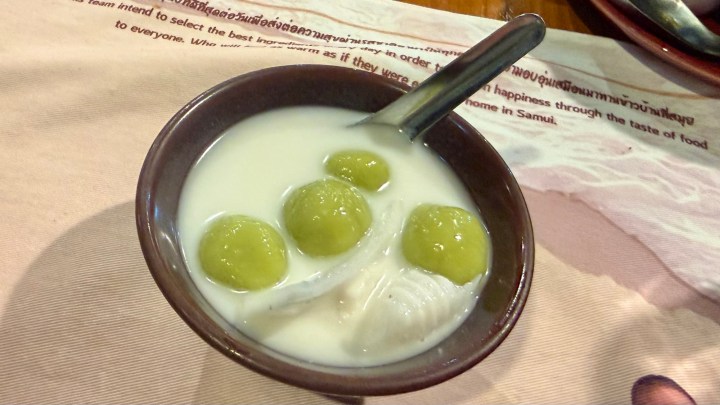 A bowl of Kanom Co, a Thai dessert made of glutinous rice flour balls filled with caramelized palm sugar, served in sweet coconut milk with fresh coconut slices.