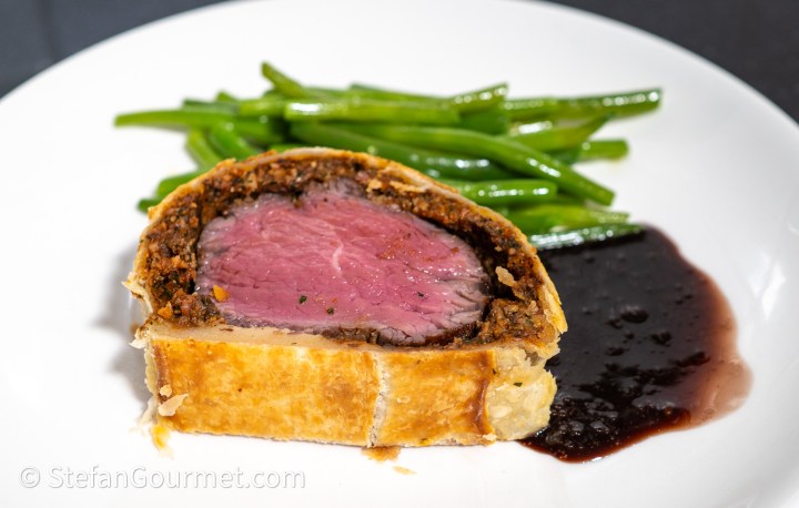 A slice of beef Wellington featuring tender pink beef inside a golden pastry crust, accompanied by a side of green beans and a dark sauce on the plate.