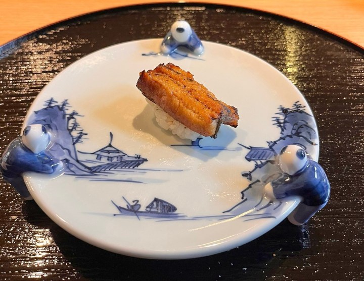 A piece of grilled eel sushi served on a decorative blue and white plate with small figurines.