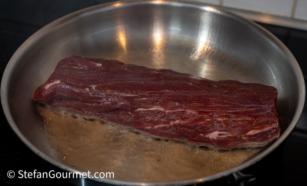 A raw piece of meat searing in a stainless steel frying pan on a stovetop.