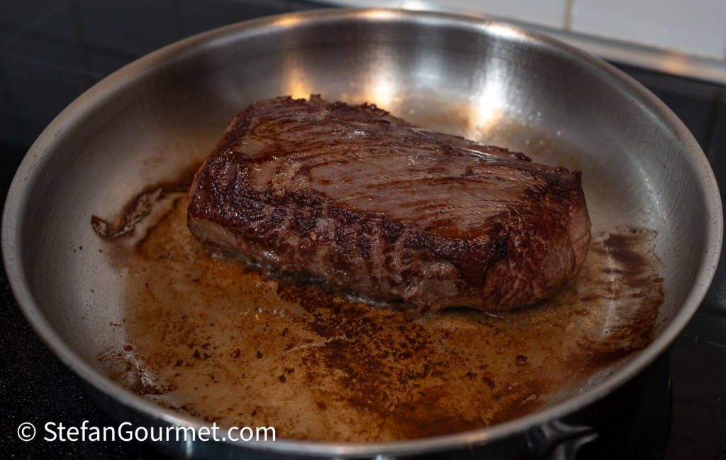 A large piece of beef steak searing in a stainless steel frying pan with brown juices.