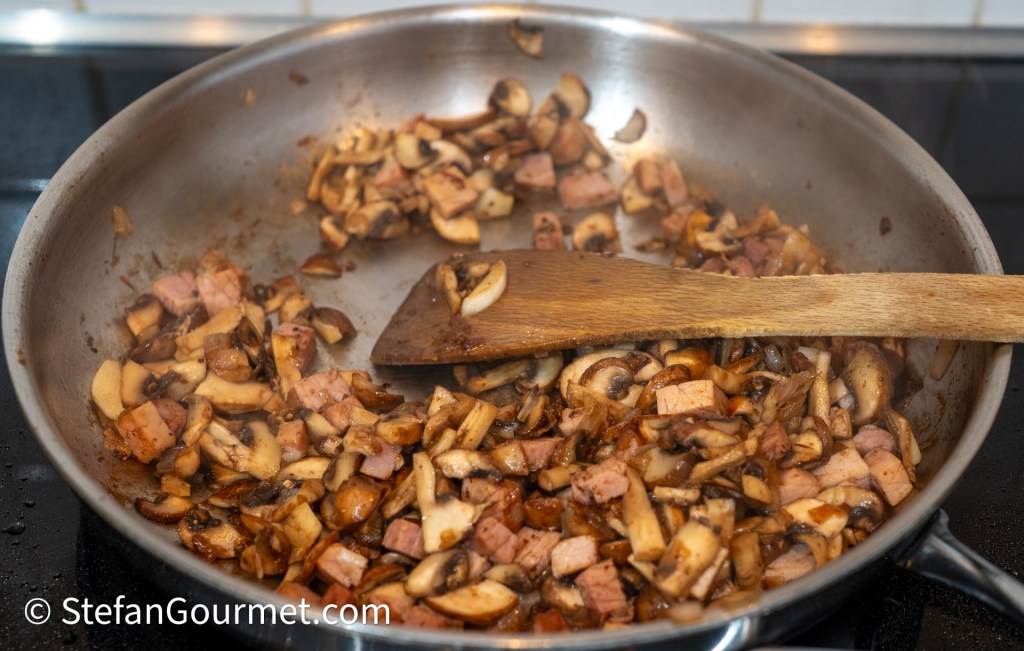 A stainless steel frying pan with sautéed mushrooms and diced ham, stirred with a wooden spatula.
