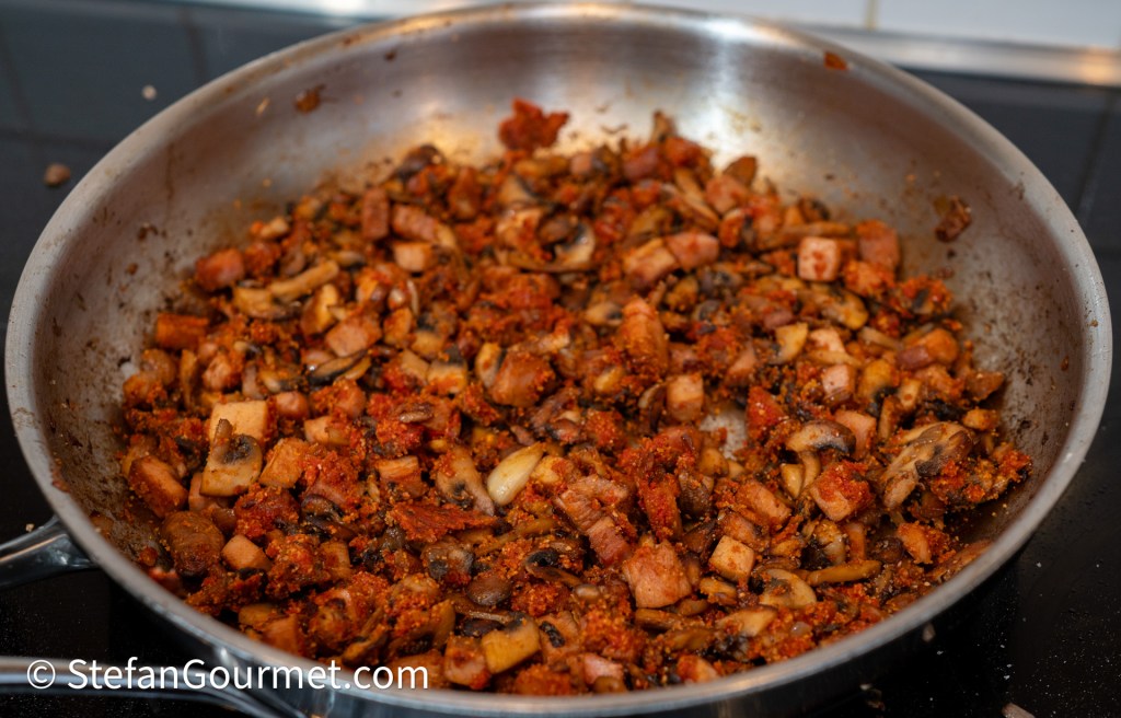 A close-up view of a stainless steel pan filled with a mixture of sautéed diced vegetables, including mushrooms and small pieces of meat, seasoned with spices.