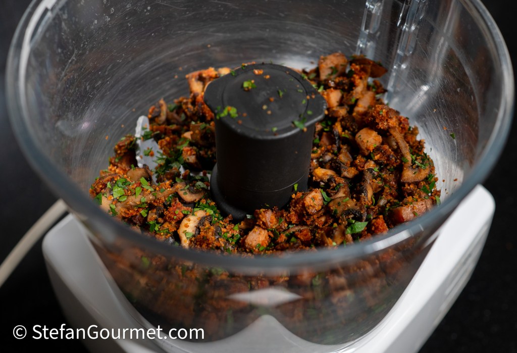A close-up view of a food processor containing a mixture of chopped herbs and vegetables, including mushrooms and textured ingredients.