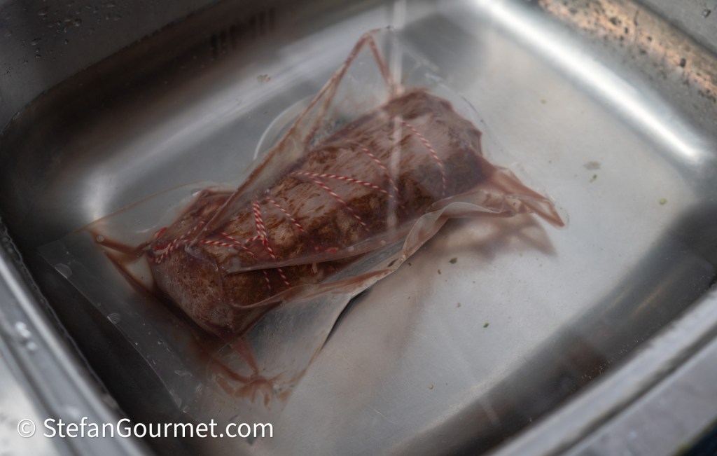 A vacuum-sealed package containing a piece of meat submerged in water in a stainless steel sink.