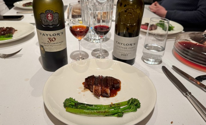 A gourmet plate featuring a piece of meat in a rich sauce, accompanied by green vegetables, with two bottles of Taylor's Port wine and glasses of wine on a dining table.