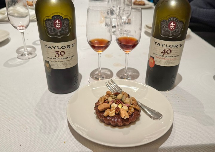A dessert tart topped with mixed nuts sits on a white plate, next to two glasses filled with tawny port wine. Two bottles of Taylor's tawny port, one labeled 30 years old and the other 40 years old, are displayed in the background.